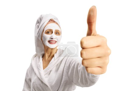 Téléchargez les photos : Woman in a bathrobe with a face mask gesturing thumbs up isolated on white background - en image libre de droit
