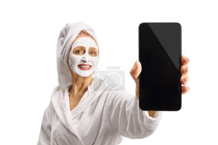 Téléchargez les photos : Woman in a bathrobe with a face mask showing a smartphone isolated on white background - en image libre de droit