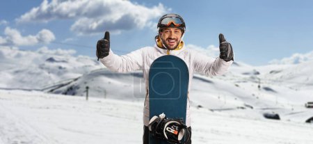 Photo for Young man with a snowboard standing on a mountain and gesturing with both thumbs up - Royalty Free Image