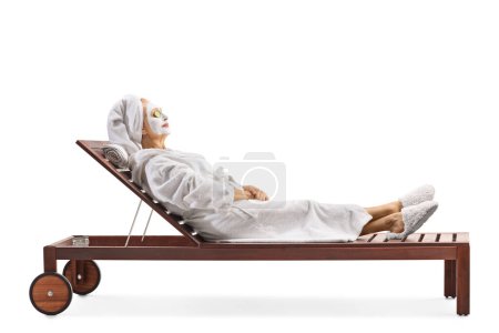 Photo for Side shot of a woman in a bathrobe and hair wrapped in towel relaxing with a white face mask on a lounge chair isolated on white background - Royalty Free Image
