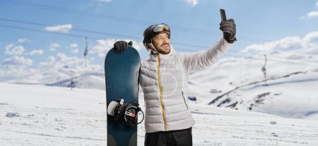 Photo for Man with a snowboard taking a selfie with a smartphone on a snowy mountain - Royalty Free Image