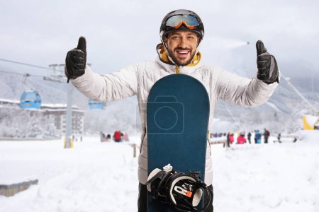 Téléchargez les photos : Young man with a snowboard gesturing with both thumbs up at a ski resort - en image libre de droit
