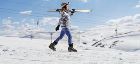 Foto de Full length profile shot of a young female with carrying skis and walking on a snowy hill - Imagen libre de derechos