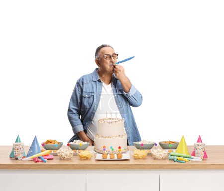 Photo for Casual mature man blowing a party horn behind a counter with cake isolated on white background - Royalty Free Image
