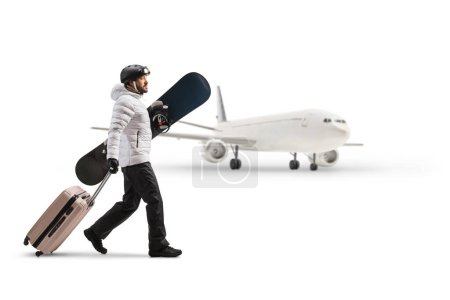 Téléchargez les photos : Young man holding a snowboard and pulling a suitcase in front of an airplane isolated on white background - en image libre de droit