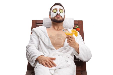 Foto de Young man with a face mask and cucumbers over eyes enjoying with a glass of cocktail isolated on white background - Imagen libre de derechos