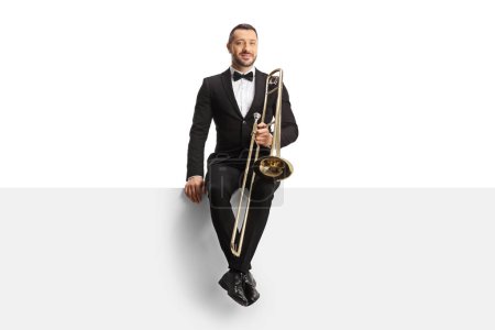 Photo for Male musician in a black suit and bow-tie sitting on a blank panel with a trombone isolated on white background - Royalty Free Image