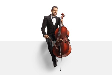 Foto de Full length portrait of a musician in a black suit and bow-tie sitting on a white panel with a cello isolated on white background - Imagen libre de derechos
