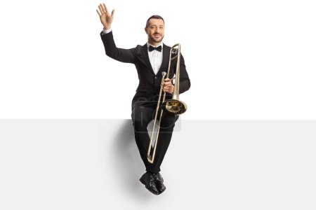 Foto de Male musician sitting on a blank panel with a trombone and waving isolated on white background - Imagen libre de derechos