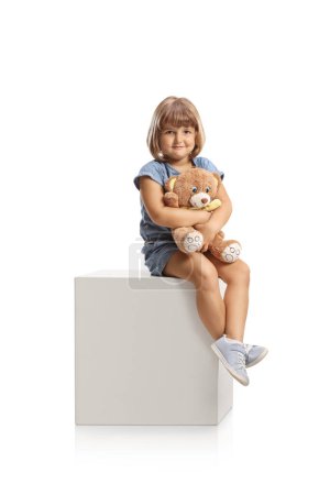Téléchargez les photos : Cute little girl holding a teddy bear and sitting on a white cube isolated on white background - en image libre de droit