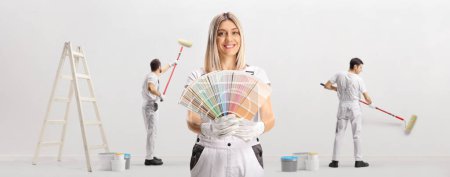 Téléchargez les photos : Female house decorator holding a color swatch palette and other workers painting a wall - en image libre de droit