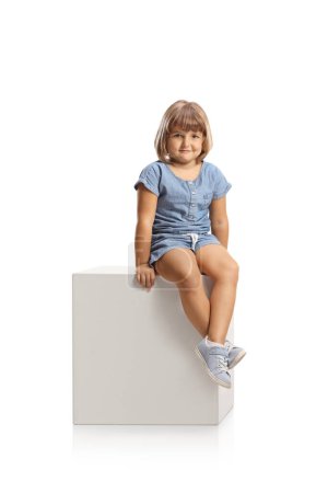 Foto de Cute little girl sitting on a white cube and smiling isolated on white background - Imagen libre de derechos