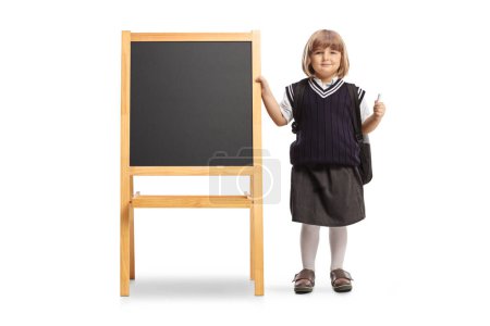 Photo for Little girl in a school uniform holding a chalk and standing next to a school blackboard isolated on white background - Royalty Free Image