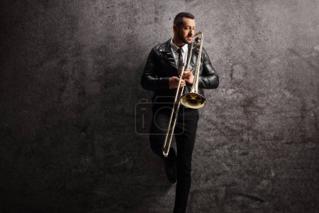 Photo for Young man holding a trombone and standing against a gray wall - Royalty Free Image