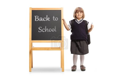 Photo for Female pupil in a school uniform holding a chalk and standing next to a blackboard with text back to school isolated on white background - Royalty Free Image