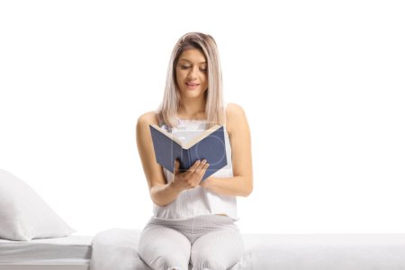 Foto de Woman in pajamas sitting on a bed and reading a book isolated on white background - Imagen libre de derechos