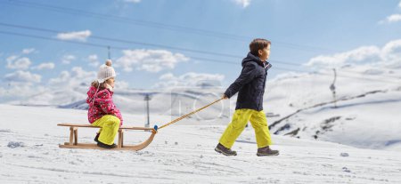 Foto de Full length profile shot of a boy pulling girl on a wooden sleigh on a snowy mountain - Imagen libre de derechos
