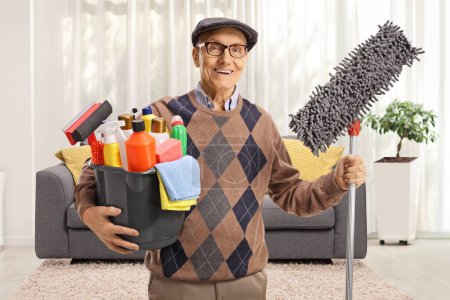 Photo for Elderly man holding a bucket with cleaning supplies and a floor mop in a living room - Royalty Free Image