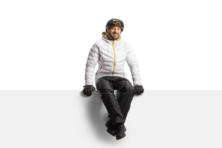 Foto de Young man wearing a skiing equipment and sitting on a blank panel isolated on white background - Imagen libre de derechos