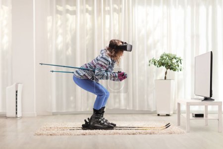 Téléchargez les photos : Full length profile shot of a young woman skiing and wearing a virtual reality headset in front of tv at home - en image libre de droit