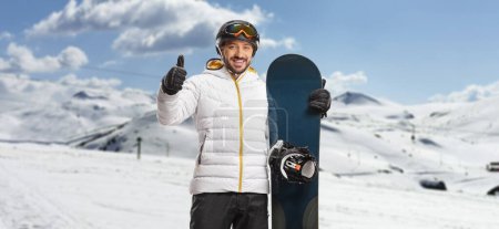 Foto de Hombre feliz con equipo de snowboard de pie en una montaña y mostrando los pulgares hacia arriba - Imagen libre de derechos