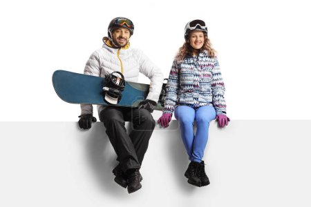 Foto de Young man and woman wearing winter equipment sitting on a blank panel and holding a snowboard isolated on white background - Imagen libre de derechos