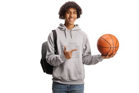 Foto de Sonriente estudiante afroamericano con una mochila sosteniendo una pelota de baloncesto y señalando aislado sobre fondo blanco - Imagen libre de derechos