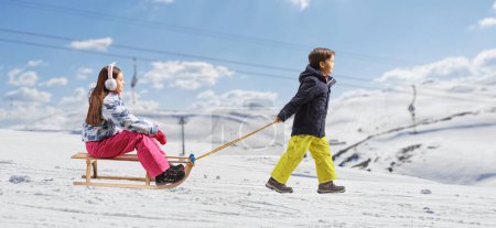 Téléchargez les photos : Full length profile shot of a boy walking and pulling a girl with a wooden sleigh on a mountain hill - en image libre de droit