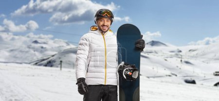 Téléchargez les photos : Homme avec lunettes et casque tenant un snowboard sur une montagne et souriant à la caméra - en image libre de droit