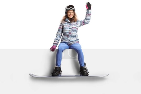 Téléchargez les photos : Jeune femme avec lunettes et casque et un snowboard assis sur un panneau blanc et agitant isolé sur fond blanc - en image libre de droit