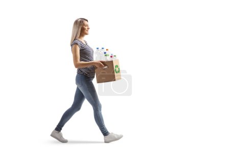 Téléchargez les photos : Full length profile shot of a young woman carrying a cardboard box with recycling plastic bottles and walking isolated on white background - en image libre de droit