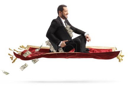 Foto de Businessman holding a briefcase with money and sitting on a flying carpet isolated on white background - Imagen libre de derechos