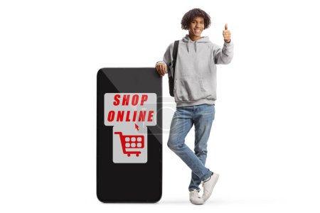 Téléchargez les photos : Young african american male gesturing thumbs up and leaning on a big mobile phone with text shop online isolated on white background - en image libre de droit