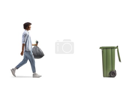 Téléchargez les photos : Full length profile shot of an african american man walking towards a bin and carrying a plastic waste bag isolated on white background - en image libre de droit