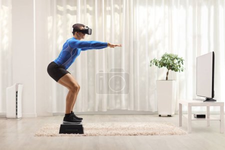 Foto de Foto de perfil completo de un hombre musculoso haciendo ejercicio con un stepper y usando un auricular vr delante de la televisión en casa - Imagen libre de derechos