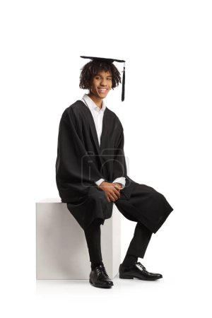 Téléchargez les photos : Homme afro-américain étudiant diplômé dans une robe assis et souriant isolé sur fond blanc - en image libre de droit