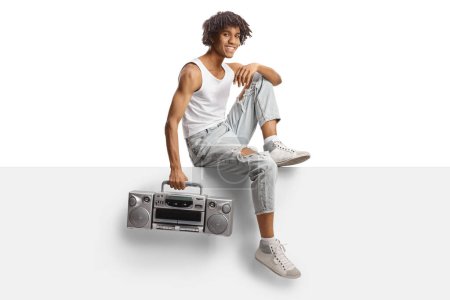 Foto de African american young man holding a boombox and sitting on a blank panel isolated on white background - Imagen libre de derechos