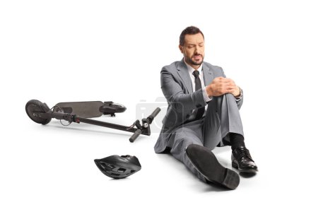 Téléchargez les photos : Homme d'affaires avec un scooter électrique assis sur le sol et tenant son genou blessé isolé sur fond blanc - en image libre de droit