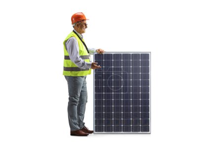 Téléchargez les photos : Mature male engineer with a vest and hardhat standing next to a photovoltaic panel isolated on white background - en image libre de droit