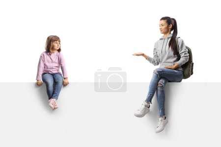 Téléchargez les photos : Female student talking to a little girl and sitting on a blank white panel isolated on white background - en image libre de droit