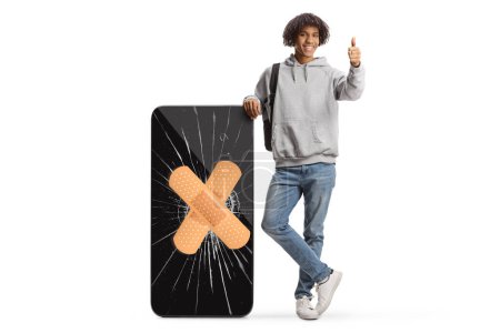 Téléchargez les photos : Young african american man leaning on a big mobile phone with cracked screen and bandage and gesturing thumbs up isolated on white background - en image libre de droit