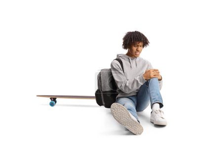 Photo for African american guy with a skateboard sitting on the ground and holding his injured knee isolated on white background - Royalty Free Image