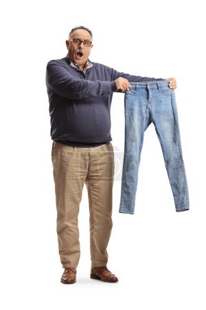 Téléchargez les photos : Portrait complet d'un homme mature surpris tenant une petite paire de jeans isolés sur le dos blanc - en image libre de droit