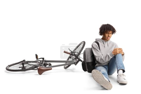 Photo for African american guy with a bicycle sitting on the ground and holding his injured knee isolated on white background - Royalty Free Image