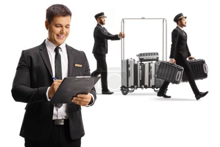 Téléchargez les photos : Réceptionniste écrivant un document et un groom poussant des valises sur un chariot à bagages isolé sur fond blanc - en image libre de droit
