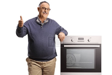 Téléchargez les photos : Smiling mature man leaning on an oven and gesturing thumbs up isolated on white backgroun - en image libre de droit