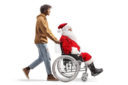 Téléchargez les photos : Full length profile shot of an african american young man pushing a santa claus in a wheelchair isolated on white background - en image libre de droit