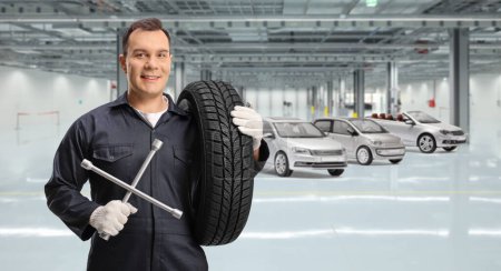 Téléchargez les photos : Mécanicien automobile portant un pneu et tenant un outil de clé à molette dans un garage de voiture - en image libre de droit
