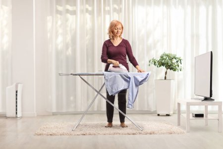 Téléchargez les photos : Mature woman ironing in front of tv in a living room - en image libre de droit