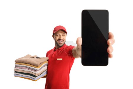 Foto de Trabajador de lavandería sosteniendo una pila de ropa doblada y mostrando un teléfono inteligente aislado en un fondo blanco - Imagen libre de derechos